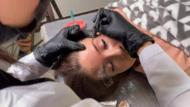 Beautician in black gloves plucks extra hairs on the model's eyebrows