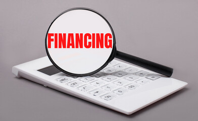 On a gray background, a white calculator and a magnifying glass with the text FINANCING. Business concept