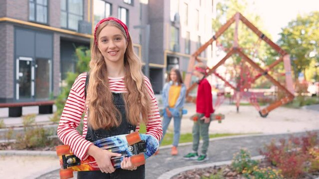 Portrait of Caucasian beautiful young stylish teen girl carrying skateboard in hands and looking at camera in good mood standing in city on street. Cute happy female kid skater outdoor Teenage concept