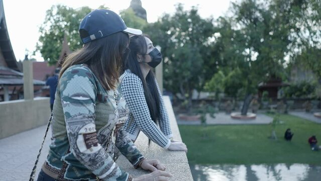 Two Thai girls in black respiratory masks lean on a stony bridge by the park and hug - slow motin 