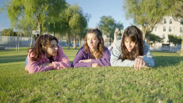 Three teenage girls lie on green grass in sunshine and chat, frontal