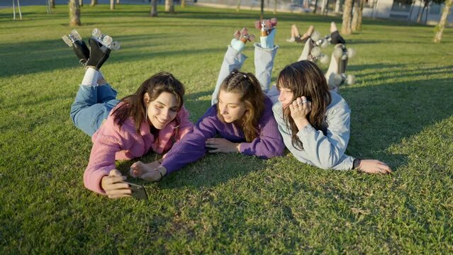 Teenage girls in colorful clothes lying on lawn take selfie with phone