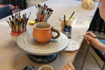 Boy coloring clay cup on a potter's wheel. Teaching pottery is Healthy free time for children. Kid painting ceramic cup on a wooden table at workshop. Hobbies, craft and handwork