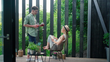 Young beautiful woman relaxing on cozy terrace, reading a book, her boyfriend is bringing her glass of wine, digital detox and weekend away concept.