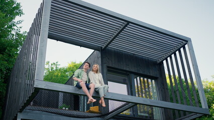 Young couple sitting and cuddling in hammock terrace in their new home in tiny house in woods,...