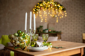 Preparing for Christmas table setting. A wooden table in a Christmas living room decorated in the trendy colors of 2022: fernwood green, venetian portico and natural linen. Candlestick, plates, wine g