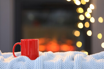Cup of hot drink on knitted blanket near fireplace at home, space for text. Cozy atmosphere