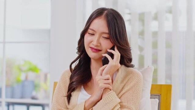 Smiling young woman talking on phone sitting at couch in the living room, happy female spending time talk to friends and family in her free time. Cheerful Asian girl having fun during conversation.