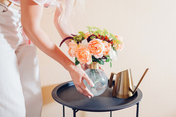 Woman puts vase with bouquet of flowers on table at home. Floral arrangement with orange roses....