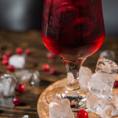 berry bright red cocktail in a large transparent glass close up, frozen berries ice inside, berries and ice are scattered around on a wooden table with free space for you text