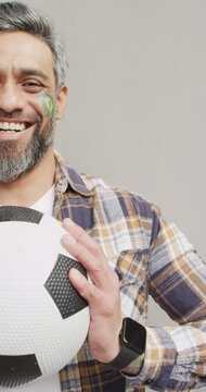 Vertical video of happy biracial man holding football with flag of brazil on cheek