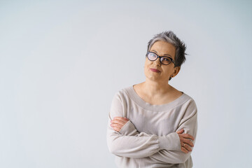 Serious slightly smiling mature grey hair woman in glasses posing with hands folded looking at camera wearing white blouse, copy space isolated on white background. Healthcare, aged beauty concept
