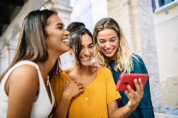 Happy young women having fun together watching funny media content on mobile phone while standing together in city street - Technology and friendship concept