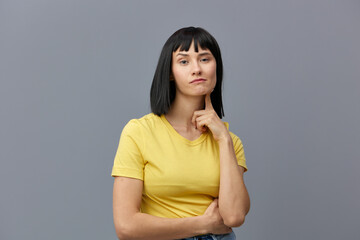 a beautiful, attractive playful woman poses standing on a gray background in a yellow T-shirt and playfully looking at the camera with her index finger touching her face