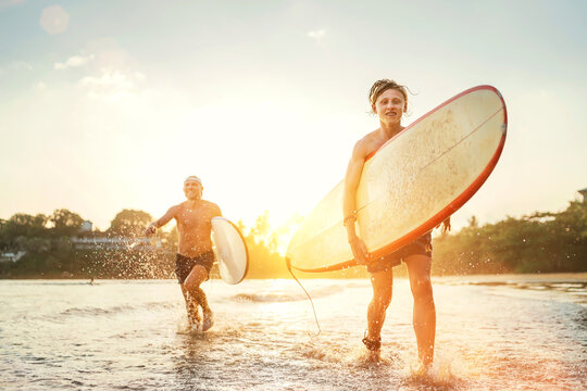 Young teen boy with a surfboard running with his father by ocean sandy beach after surfing. They are smiling and enjoying a beautiful sunset light. Family active vacation concept..