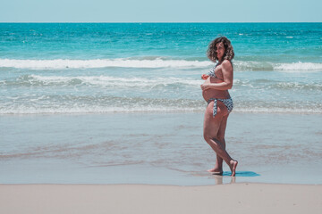 Happy pregnant woman on the beach. High risk pregnant adult lady smiles on the beach. Miss in a bikini with a big fat belly of eight months. Overweight and health concept enjoying her unborn baby 