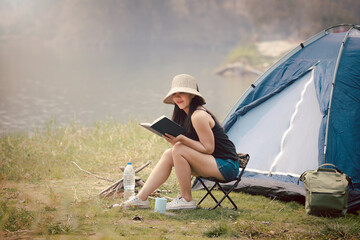 Happy woman sitting on the camping chair and reading a book in front of the camping tent at meadow near lake. Recreation and journey outdoor activity lifestyle. Travel and adventure theme.