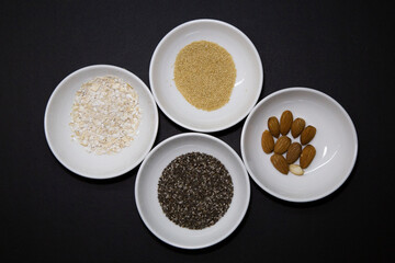 Almonds, chia and other healthy vegan seeds in 4 round white porcelain bowls on a black background