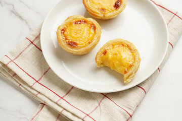 Traditional portuguese vanilla pudding puff pastry pastel de nata on white plate on checkered kitchen towel with a cup of coffee in white porcelain