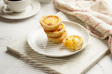 Traditional portuguese vanilla pudding puff pastry pastel de nata stacked on each other on white plate on marble board with a cup of coffee in white porcelain