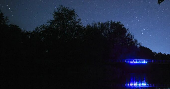 The movement of stars in the night sky over the bridge with colored backlighting, timelapse of the night landscape. Silhouette of trees.