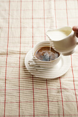 Pouring milk into white porcelain cup with black coffee on checkered table cloth