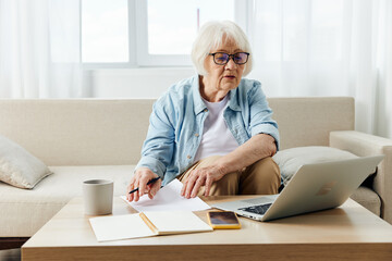a pleasant elderly lady, passionate about work, sits in her apartment on a comfortable sofa in stylish clothes and uses a laptop and looks at her notebook for notes