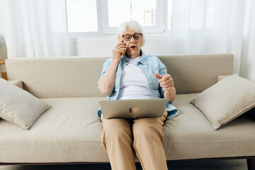 a cute elderly woman is sitting at home working on a laptop resting on the couch and talking on the phone actively gesturing with her hand