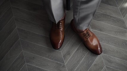 Top view of a man's leg in classic brown leather brown men's shoes. Man in brown shoes and grey trousers stands on grey parquet. Close up