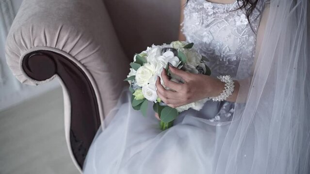 Bride in white wedding dress holds her wedding bouquet of peonies, roses and eust. Close-up of the hands of the bride with a bouquet in her hands, sitting on a classic gray sofa