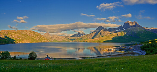Countryside landscape in Nordland, Bodo, Norway with.glistening waters on a lake near a majestic mountainscape during sunset. Blue sky with lush green grass and a little house on a river bank