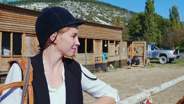 Happy female equestrienne with harness on shoulder looks at horse standing nearby. Rider caucasian girl is near stabling on paddock fenced area. Jockette smiling, resting next to palfrey outdoor
