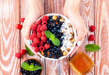 The girl's hands hold a bowl of muesli with ripe berries, yogurt and honey on the wooden table....