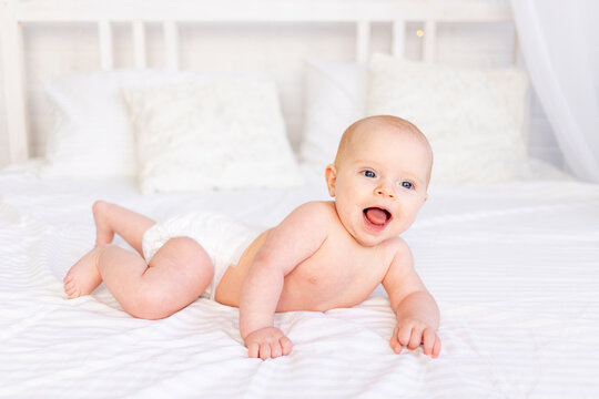 laughing baby girl in diapers in a crib on a white cotton bed lying on her stomach in the nursery smiling, newborn morning