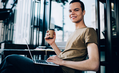 Positive student browsing laptop and drinking cup of takeaway coffee