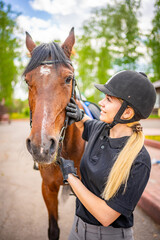 Fototapeta premium Lovely young woman wearing helmet stroking to her brown horse