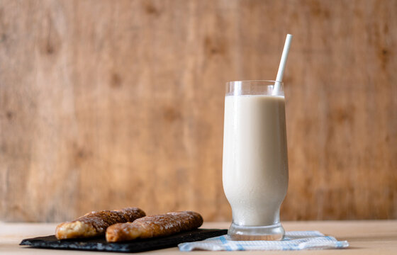 Side view. Glass with horchata, accompanied by two fartons, on a wooden background. 