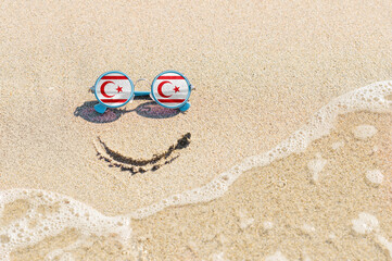 Fototapeta na wymiar Sunglasses with flag of Turkish Republic of Northern Cyprus on a sandy beach. Nearby is a sea lightning and a painted smile. The concept of a successful vacation in the resorts of North Cyprus.