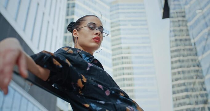Urban style seductive girl wearing colored jacket short skirt and cool futuristic glasses posing leaning back against glass parapet near high rise skyscrapers. Handsome female fashion model