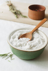 Salt in a ceramic bowl with a wooden spoon and a sprig of dry rosemary on the table. Rustic style. Natural resource. Vertical orientation.
