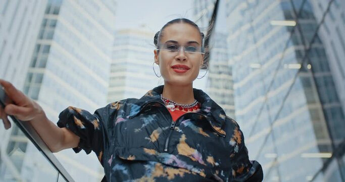Urban style seductive girl wearing colored jacket and cool futuristic glasses posing makes air kiss and smiles looking at camera standing near high skyscrapers modern buildings. Female fashion model