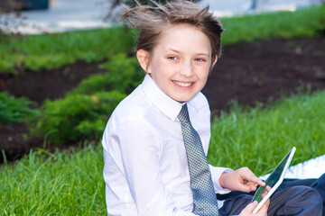 a boy in a white shirt and tie writes in a notebook outside