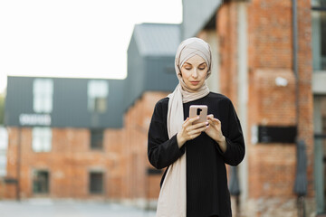 Attractive smiling muslim woman in hijab texting on smart phone standing on background of buildings in city street. Portrait of young arabian businesswoman using mobile phone in city.