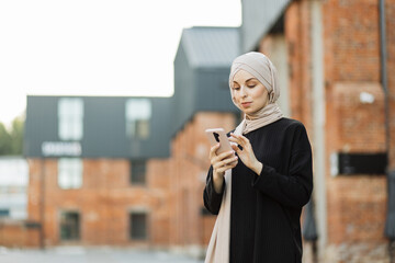 Fototapeta premium Attractive smiling muslim woman in hijab texting on smart phone standing on background of buildings in city street. Portrait of young arabian businesswoman using mobile phone in city.