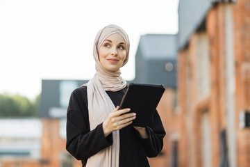 Attractive smiling muslim woman in hijab texting on tablet standing on background of buildings in...