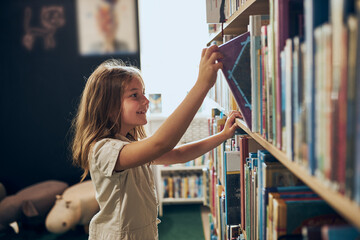 Fototapeta Schoolgirl choosing book in school library. Smart girl selecting literature for reading. Learning from books. Benefits of everyday reading. Child curiosity. Back to school obraz