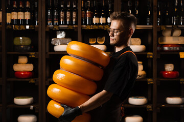 Handsome cheese sommelier with yellow wheels of cheese in shop. Worker of food cheese store...