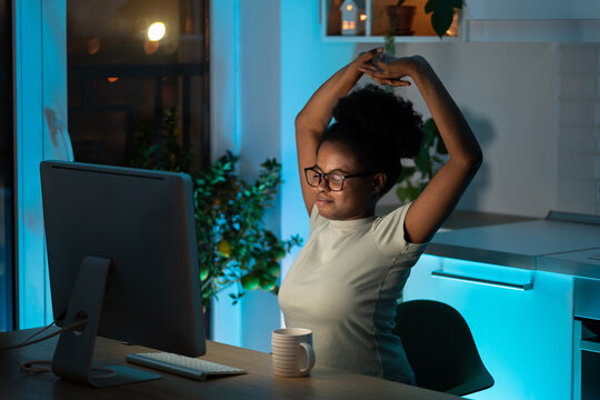 Tired African American young woman takes a break from work on computer at home office, does exercise and stretching hands up. Black student girl wants to sleep after a long work at pc late at night