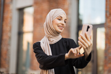 Muslim business woman talking on mobile phone outdoors. Professional woman in black suit and hijab walking on old city street checks her smartphone