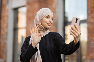 Young smiling happy muslim woman doing video call using smartphone at the city. Arabian woman blogger wearing hijab speaking on the smart phone with her audience walking in the street.